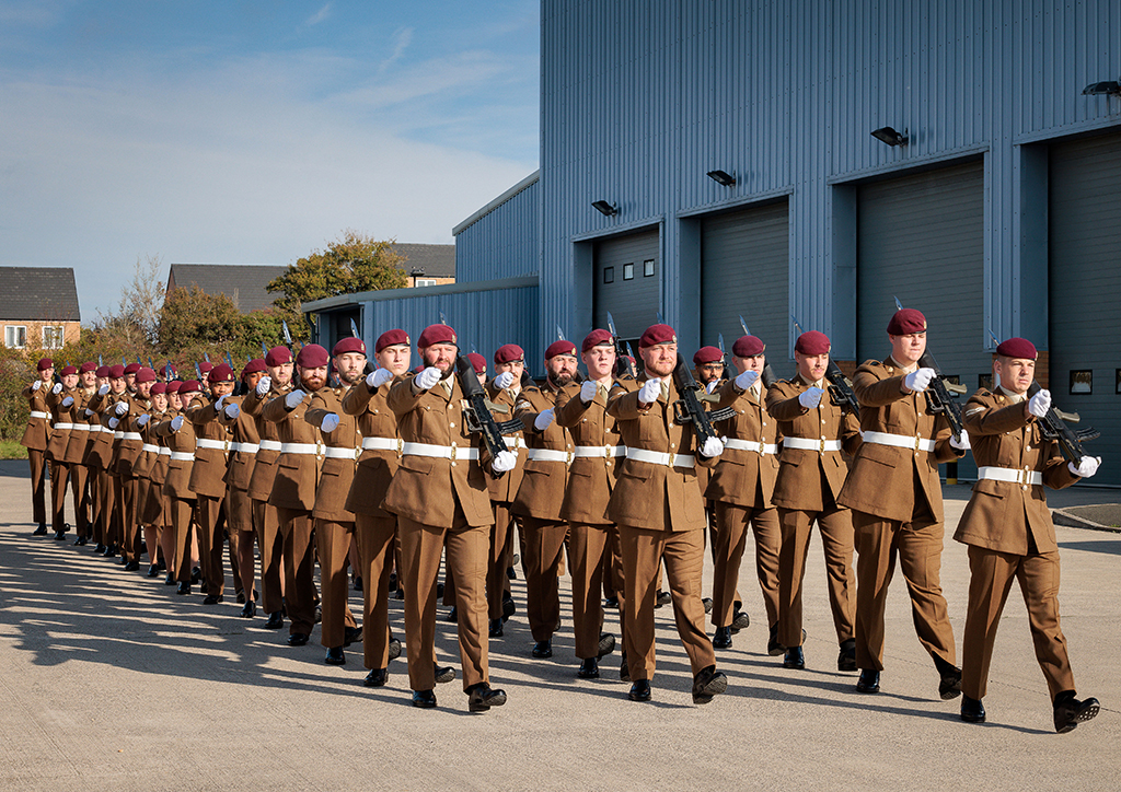 The 47 Air Despatch Squadron parade marches forward from left to right with shouldered rifles; the Squadron buildings in the background.